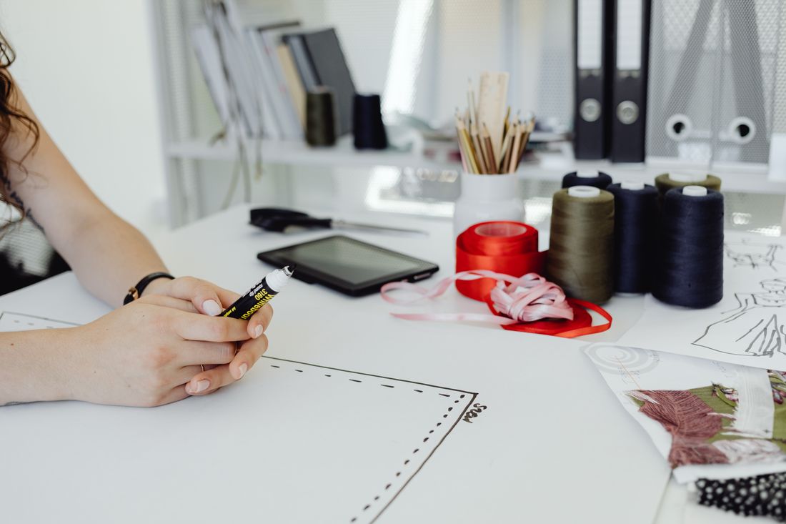 Fashion sketches, ribbon reels and drawing tools on a white desk.